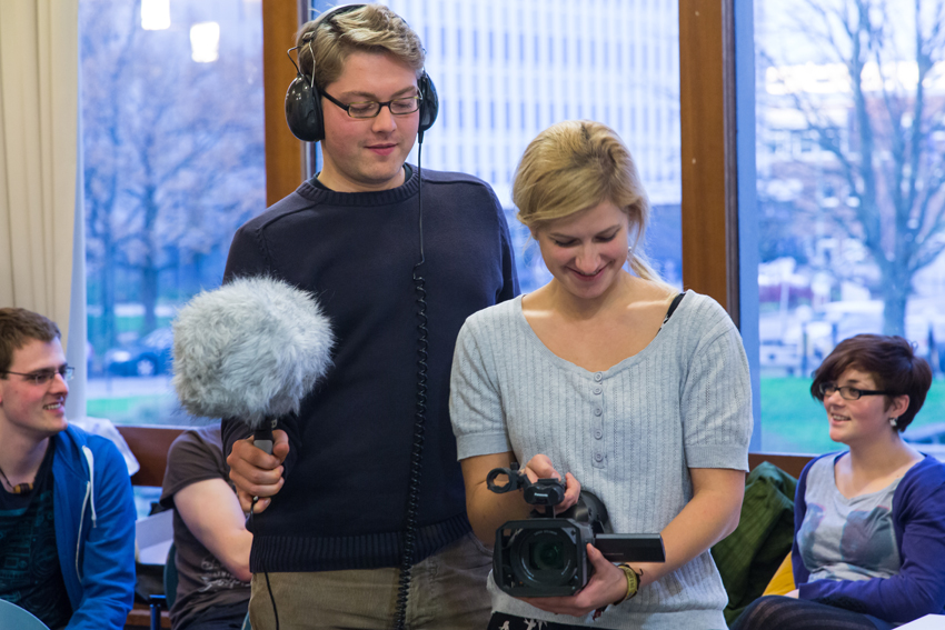 Zwei Studenten probieren das Recording-Equipment aus: ein Junge hält ein Mikrofon mit einem flauschigen Fellwindschutz, auf dem Kopf hatte einen großen Kopfhörer. Das Mädchen rechts neben ihm hält einen Videorecorder.